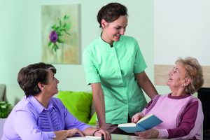 Picture includes a nurse in a care home talking to two elderly patients sat at a table reading books.