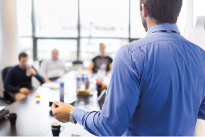 Board meeting photo depicts a man standing at the head of table presenting a speech, while people sit around a table and listen with coffees.